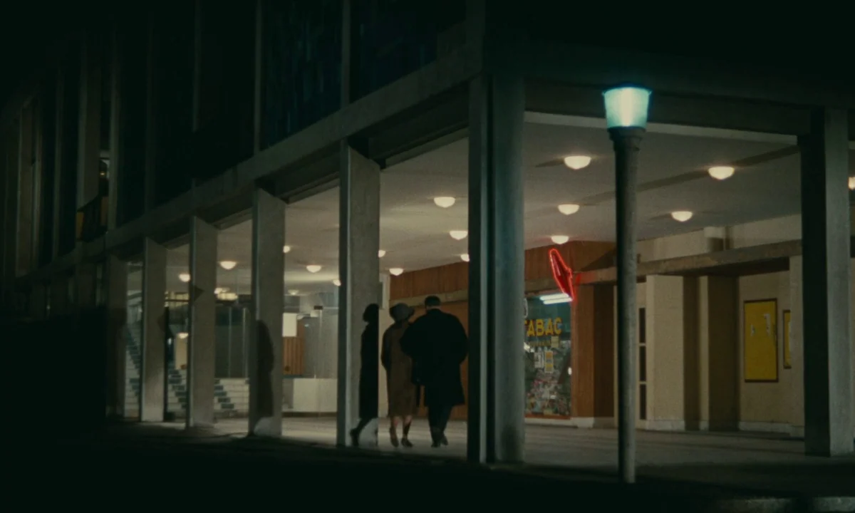 Muriel (1963) movie still: wide — A modern concrete building arcade at night features a row of lit recessed ceiling lights. A man and woman in…
