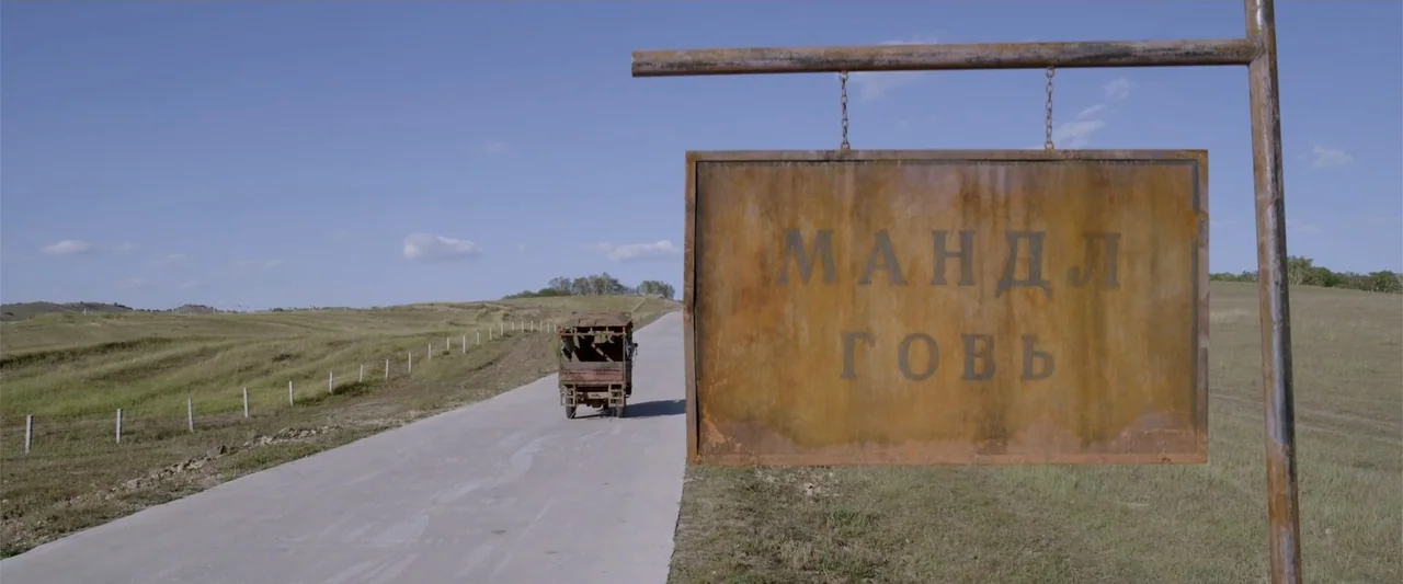 Skiptrace (2016) movie still: wide — A rusted metal sign on a post stands beside a quiet asphalt road. A small truck drives into the distance…