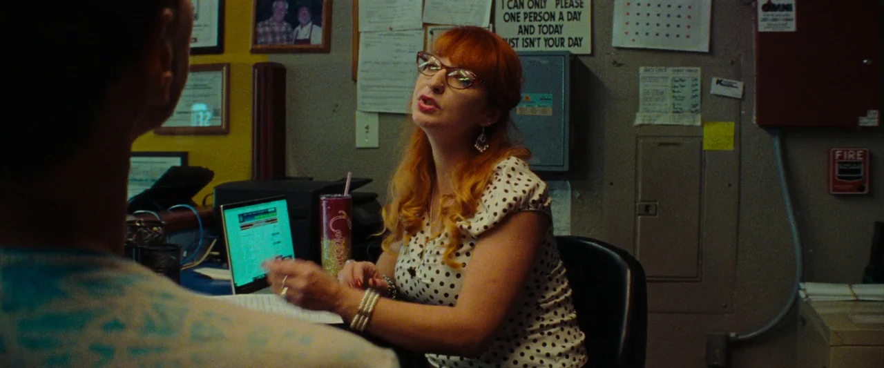 Red Rocket (2021) movie still: medium — An administrative office features cluttered walls with taped memos and a printed sign. A woman in a…