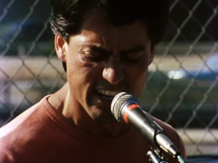 Mur Murs (1981) movie still: closeup — A man stands outdoors in front of a metal chain link fence. He is singing into a handheld microphone held…