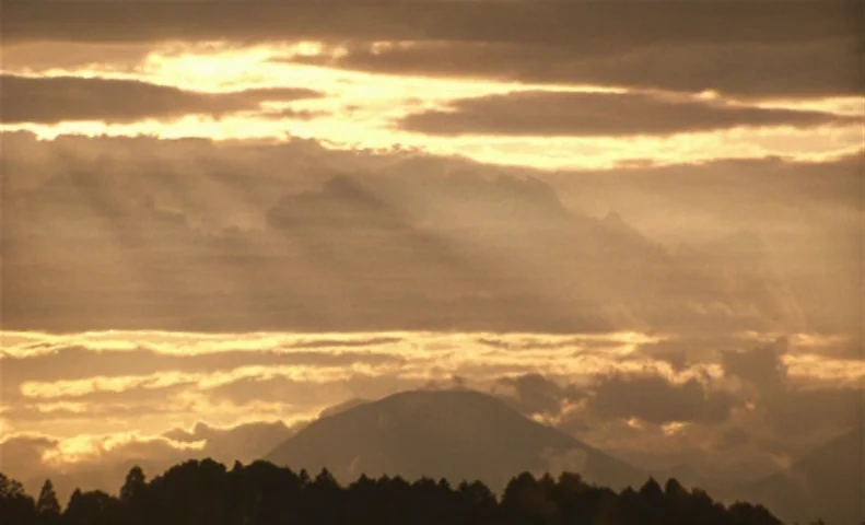 The Taste of Tea (2004) movie still: establishing — Layers of illuminated golden clouds drift above a dark, silhouetted mountain and tree line.…