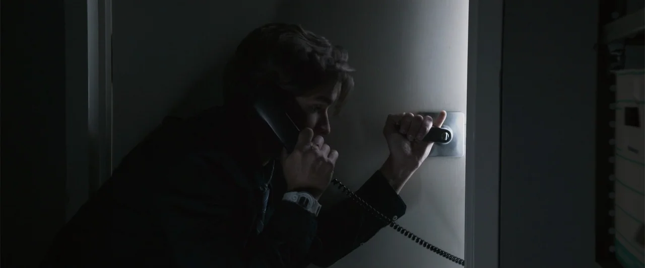 Office Uprising (2018) movie still: medium — A dimly lit indoor space with a single bright vertical light source. A young man talks on a corded…