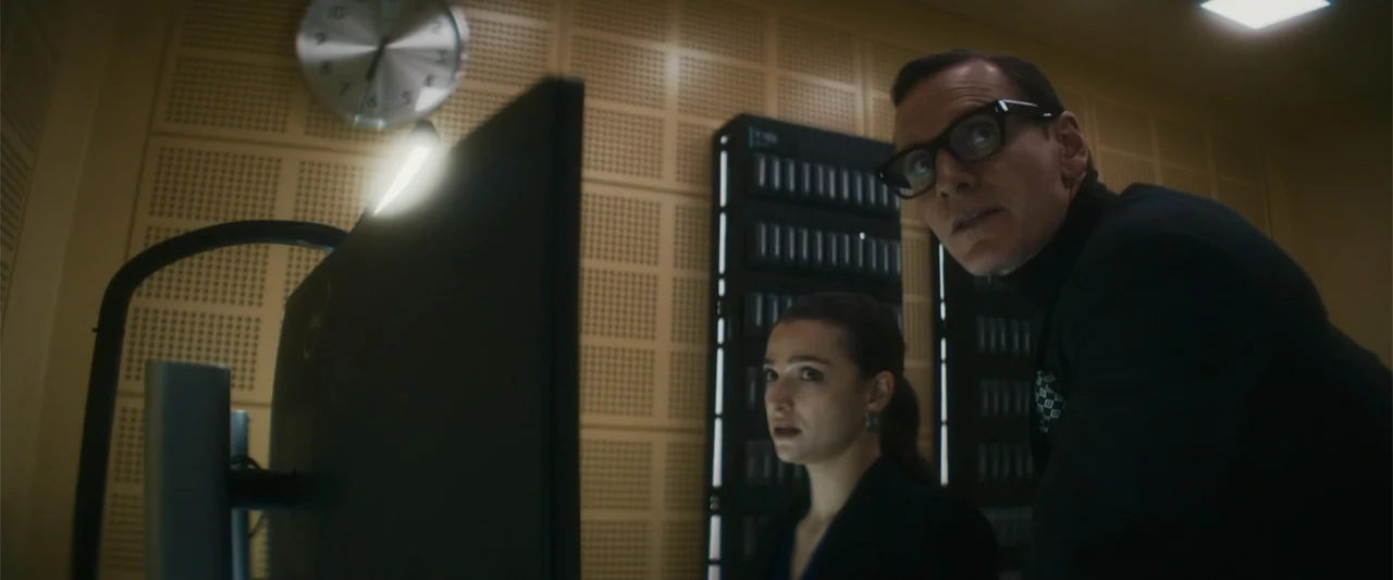 Black Bag (2025) movie still: medium — A dark server room with acoustic wall panels and a wall-mounted analog clock. A man and a woman stand before…