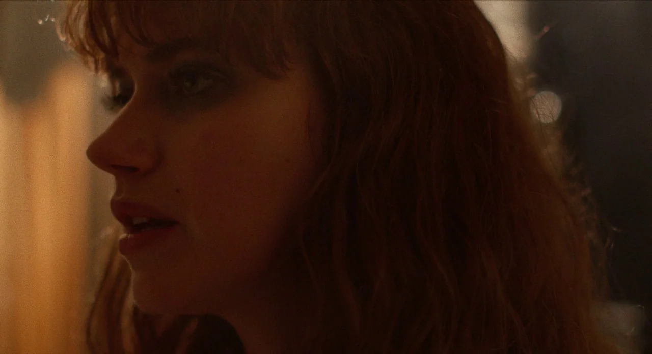 The Art of Self Defence (2019) movie still: closeup — A dimly lit indoor space with blurred background highlights. A woman with wavy hair is…