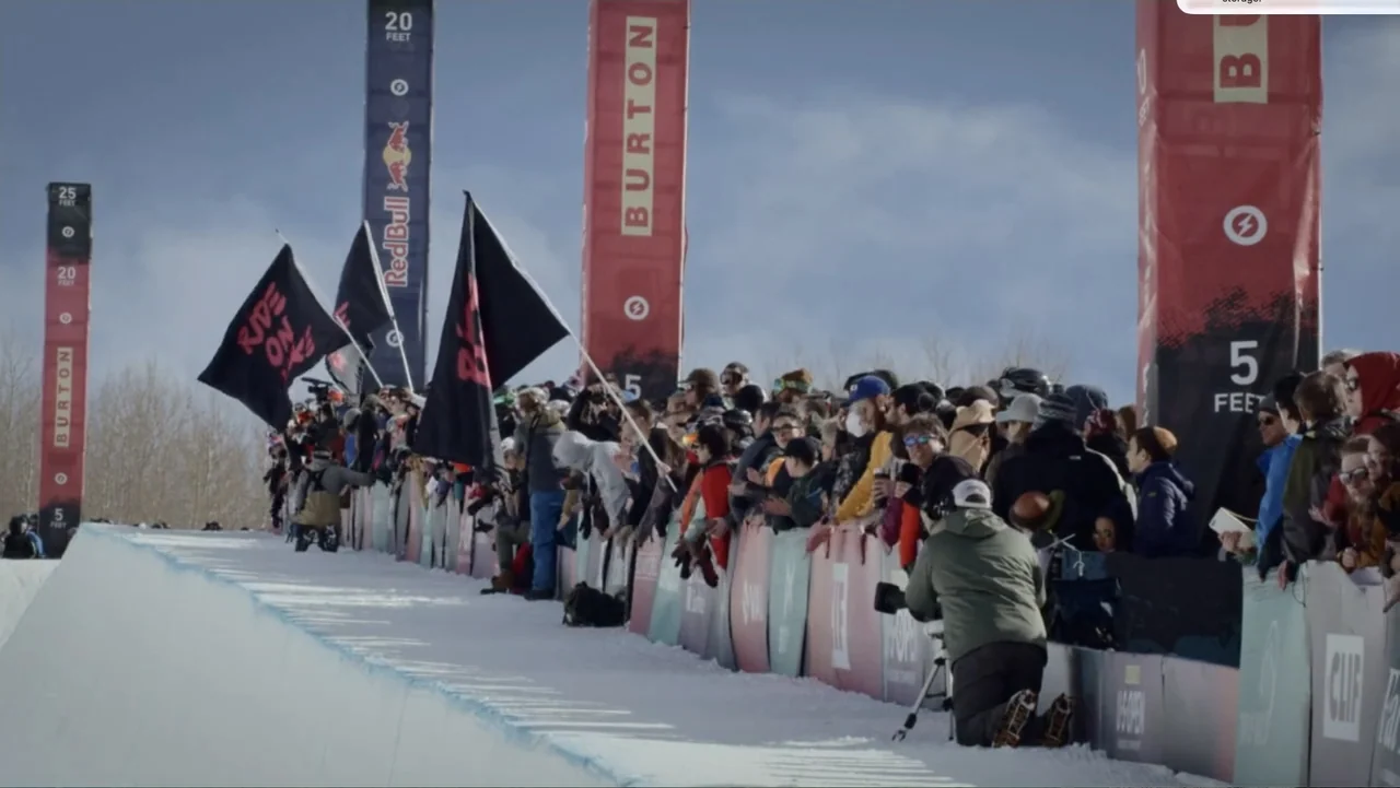 Dear Rider: The Jake Burton Story (2021) movie still: wide — A snowy outdoor arena features tall branded banners and a line of spectators watching.…