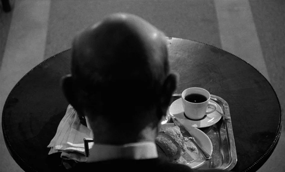 The Man From London (2007) movie still: medium — A man sits at a round wooden table topped with a silver food tray. The tray holds a small white cup…