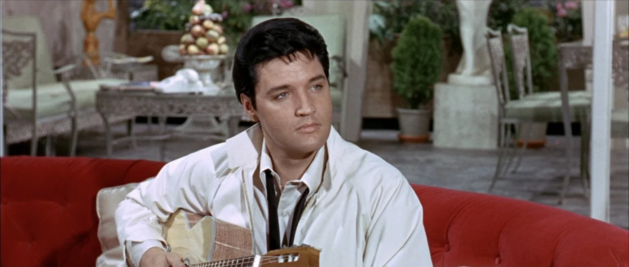 Clambake (1967) movie still: medium — A 1960s living room features a plush red sofa and outdoor patio furniture visible. A man with dark pompadour…