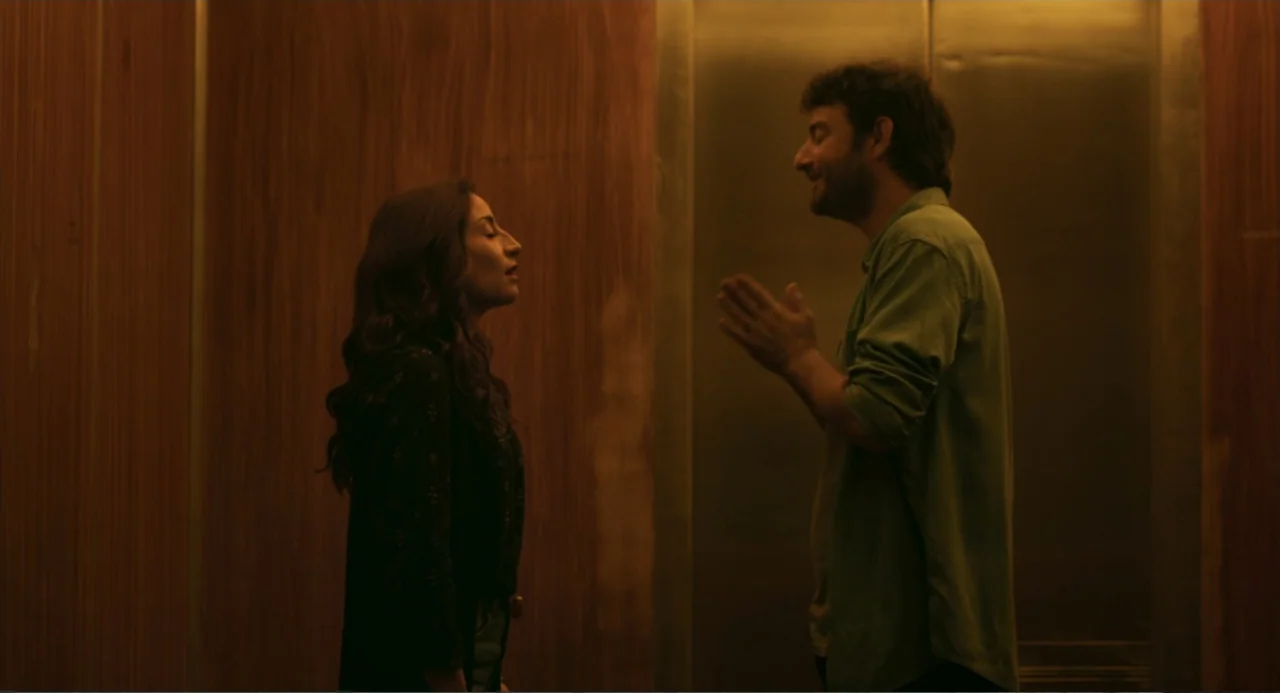 The Lift (2021) movie still: medium — An elevator interior with wood-paneled walls and metallic doors under warm artificial lighting. A woman and a…