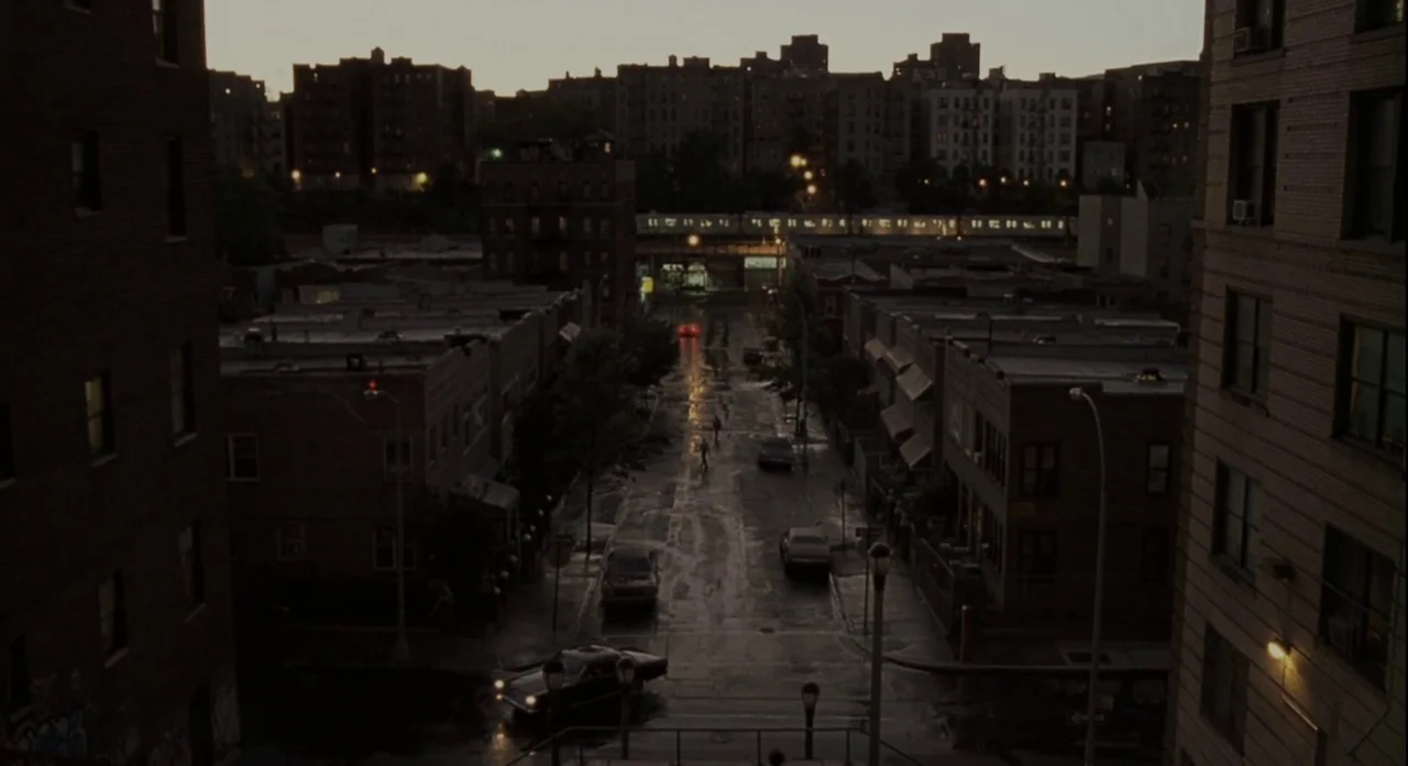 American Gangster (2007) movie still: establishing — A dense urban block is framed by tall brick apartment buildings on both sides. A train passes…