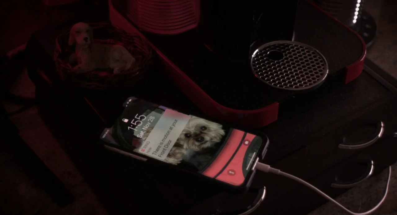 Happer’s Comet (2022) movie still: closeup — A mobile phone sits on a dark surface next to a red coffee machine. The phone screen shows a security…