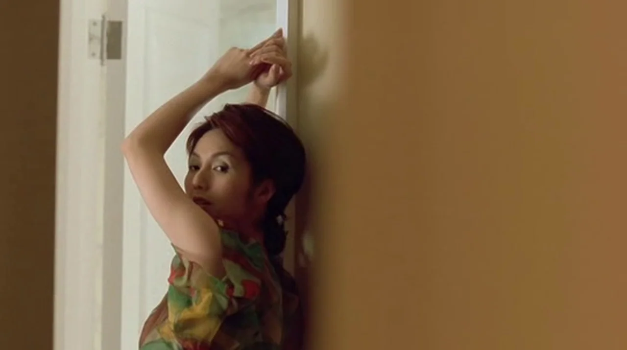 Dumplings (2004) movie still: medium — A woman stands in a bright indoor doorway adjacent to a solid tan wall. She poses with her hands lifted above…