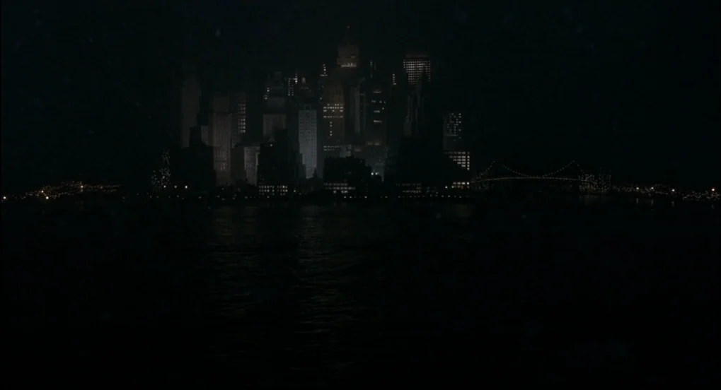 The Hudsucker Proxy (1994) movie still: establishing — A distant metropolitan skyline at night is viewed across a dark, reflective harbor. Several…