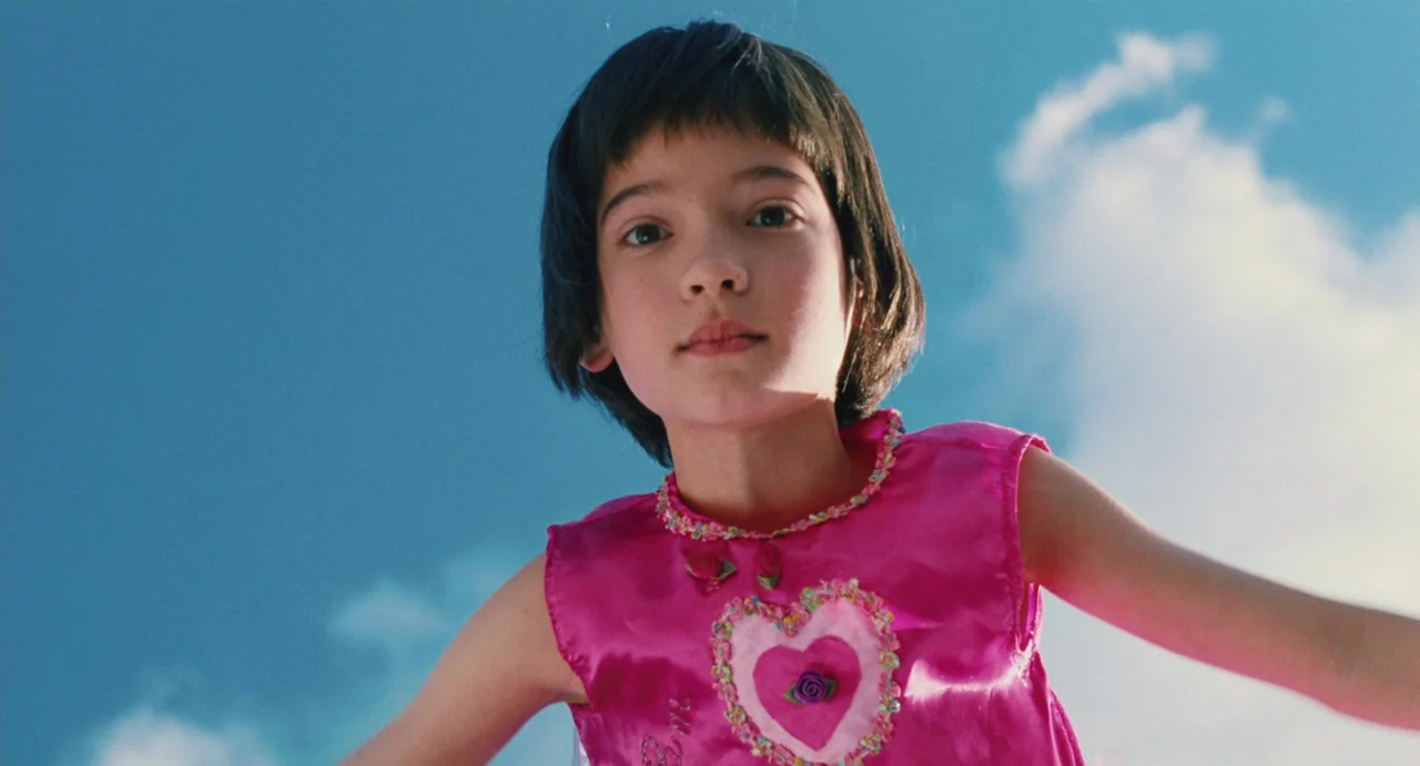 Ma Vie En Rose (1997) movie still: closeup — A young child is positioned against a bright blue sky filled with white clouds. She looks down at the…