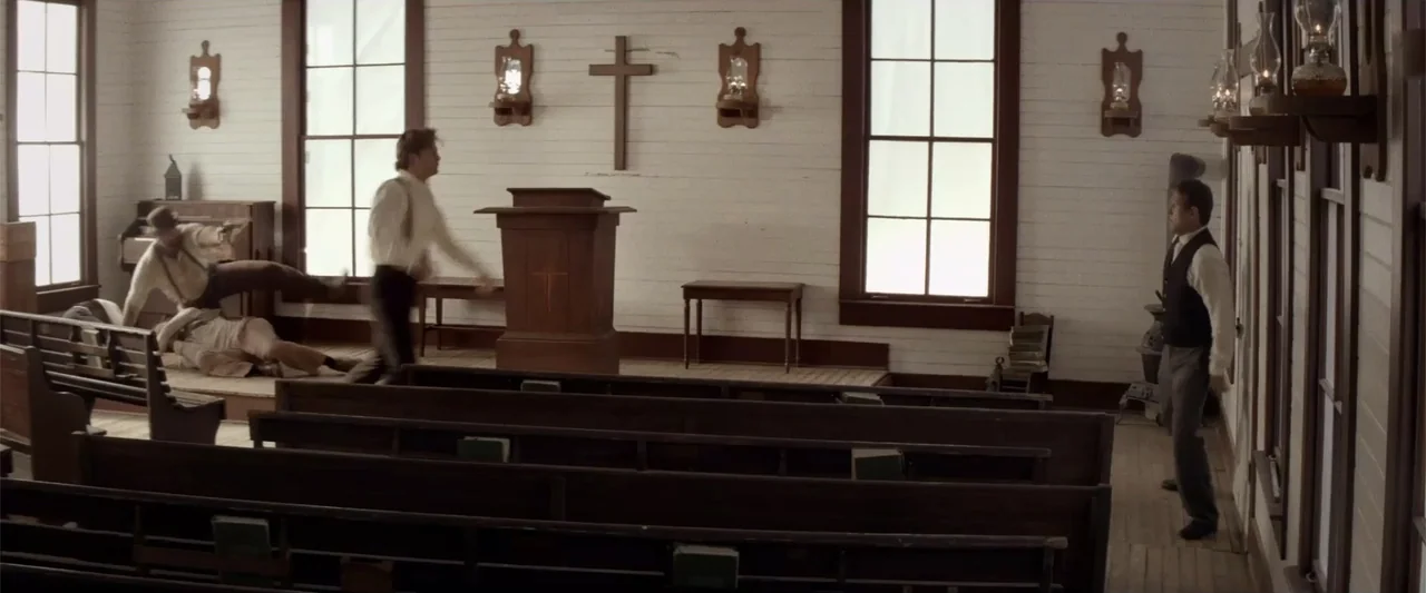 Matar o morir (1984) movie still: wide — A rustic wooden church with white horizontal siding and a central wooden pulpit. Three men engage in a…
