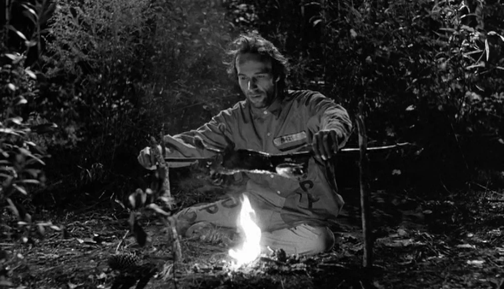 Down By Law (1986) movie still: medium — A man in a prisoner uniform sits in a dark clearing at night. He turns a spit over a small fire to cook meat.