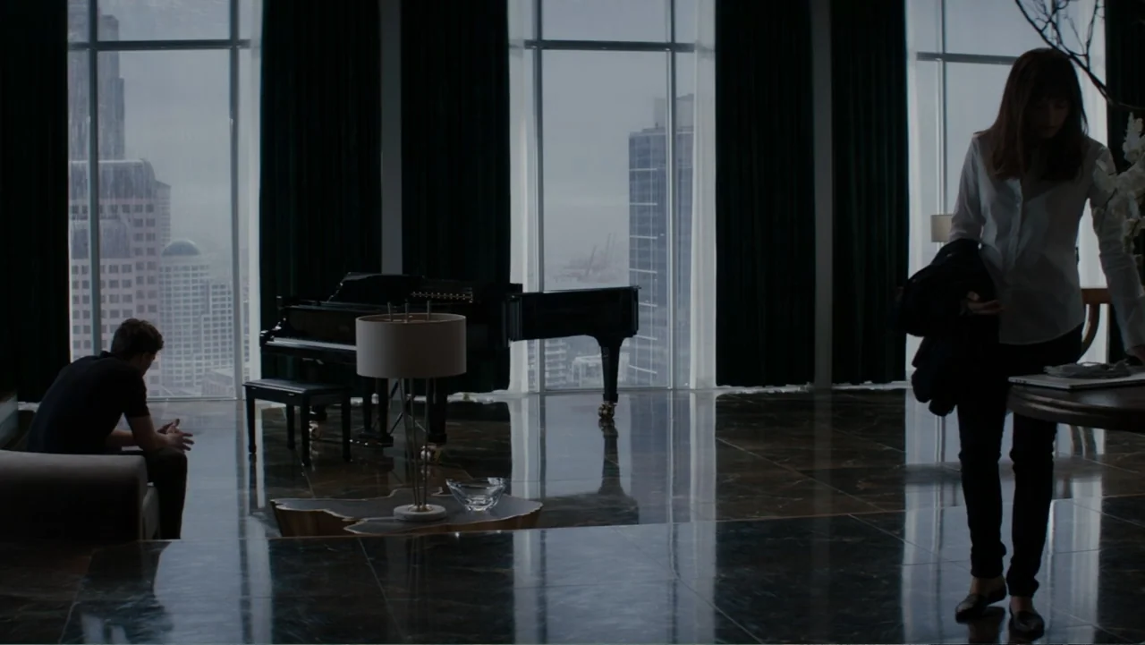 Fifty Shades of Grey (2015) movie still: wide — A spacious high-rise apartment overlooks a city skyline through large windows. A man sits on a couch…