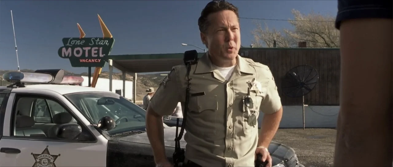 Joy Ride (2001) movie still: medium — A law enforcement officer in a tan uniform stands before a motel parking lot. He speaks to a person whose…