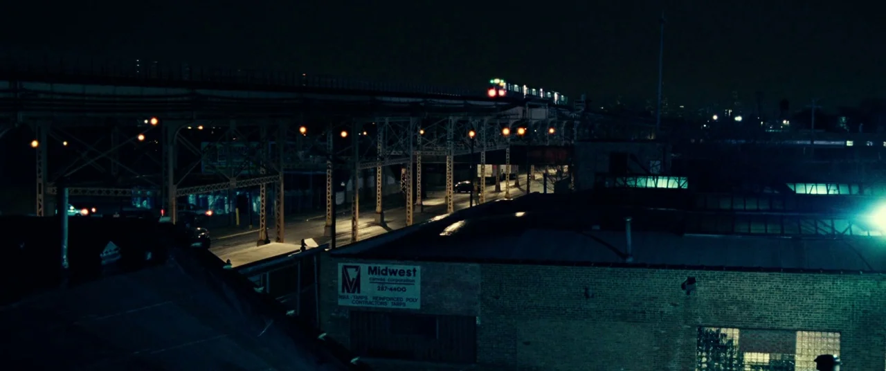 Widows (2018) movie still: establishing — An urban industrial area at night with a brick building and elevated railway tracks. A passing train glows…