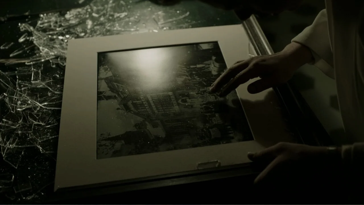 A Cure for Wellness (2017) movie still: closeup — A dark workspace features broken glass shards scattered across a surface beside a photograph. A…