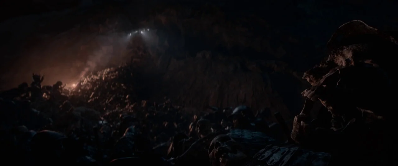 Tomb Raider (2018) movie still: wide — A dark subterranean cavern contains a massive mound of skeletal remains and armor. Sparse light highlights…