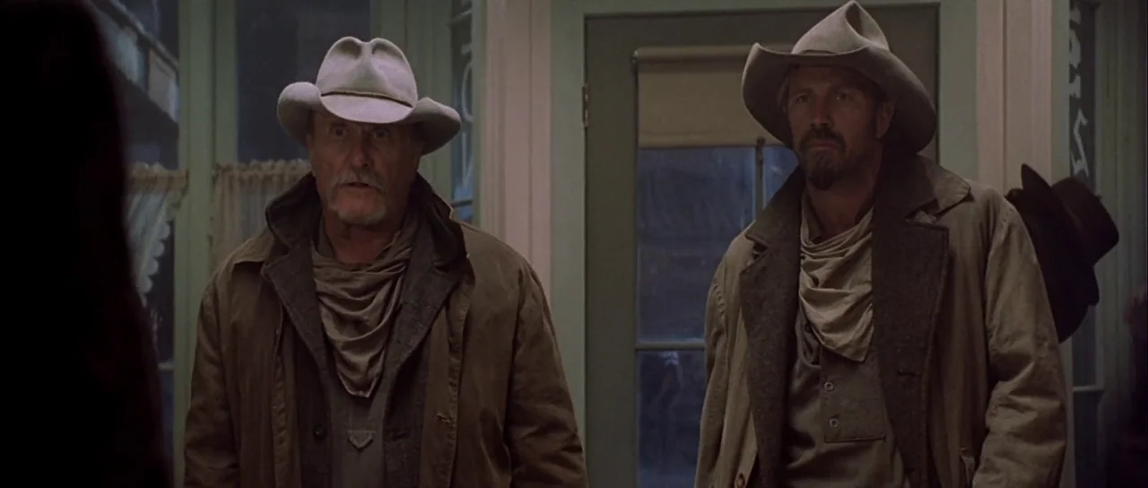 Open Range (2003) movie still: medium — A dim interior space features pale walls and a central door with glass panes. Two men wearing western-style…