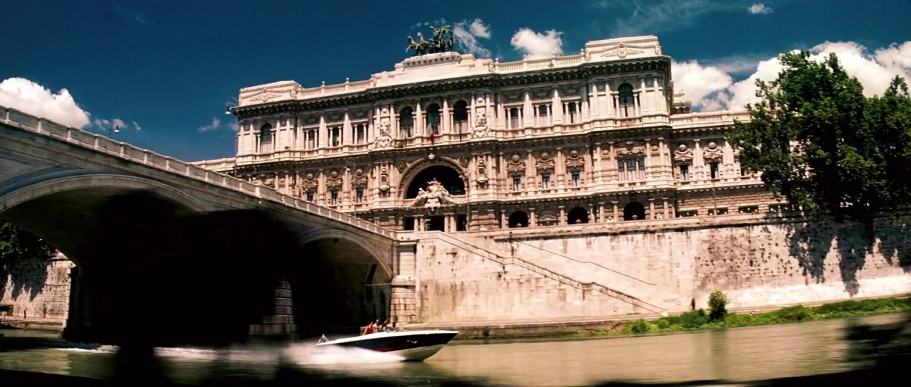 Down With Love (2003) movie still: wide — A massive historic stone building sits prominently behind a river bridge on a sunny day. A small speedboat…