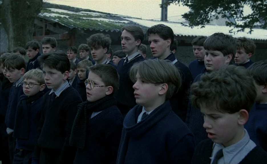 Au Revoir Les Enfants (1987) movie still: medium — A group of boys in matching navy sweaters stands outdoors on a grey day. They are arranged in…