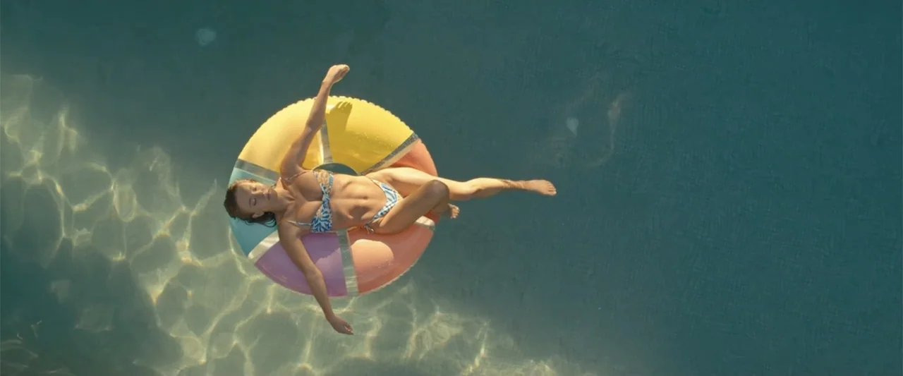 Anyone But You (2023) movie still: fullbody — A clear blue swimming pool contains a bright multi-colored circular inflatable ring. A woman in a…