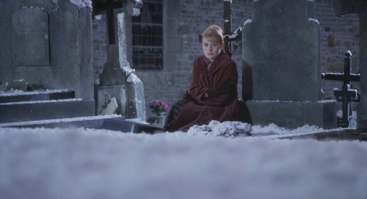 Populaire (2012) movie still: medium — A snow-covered graveyard at night featuring weathered stone tombs and monuments. A young woman sits in the…