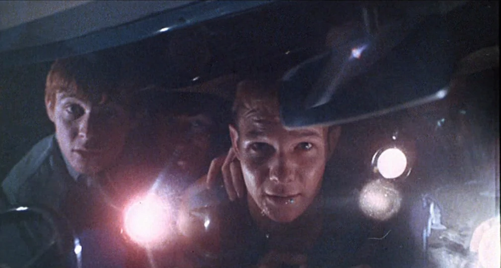 Midnight Cowboy (1969) movie still: medium — Two men sit inside a vehicle at night while scanning the darkness ahead. They hold bright flashlights…