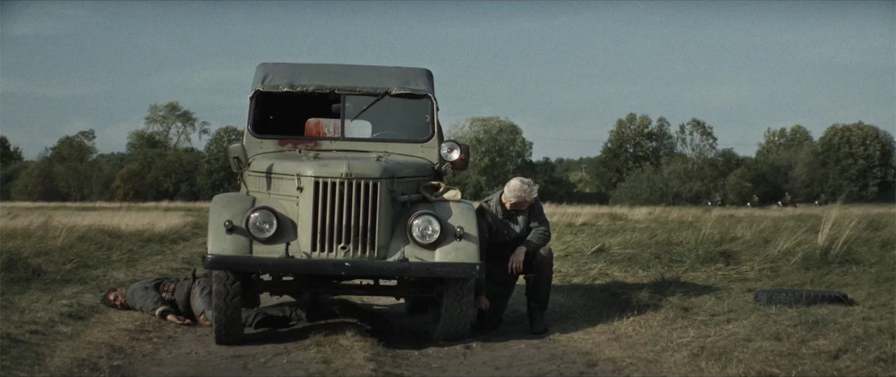 Sisu: Road to Revenge (2025) movie still: wide — A wide, flat field surrounds a vintage olive drab military vehicle parked on dirt. An older man…