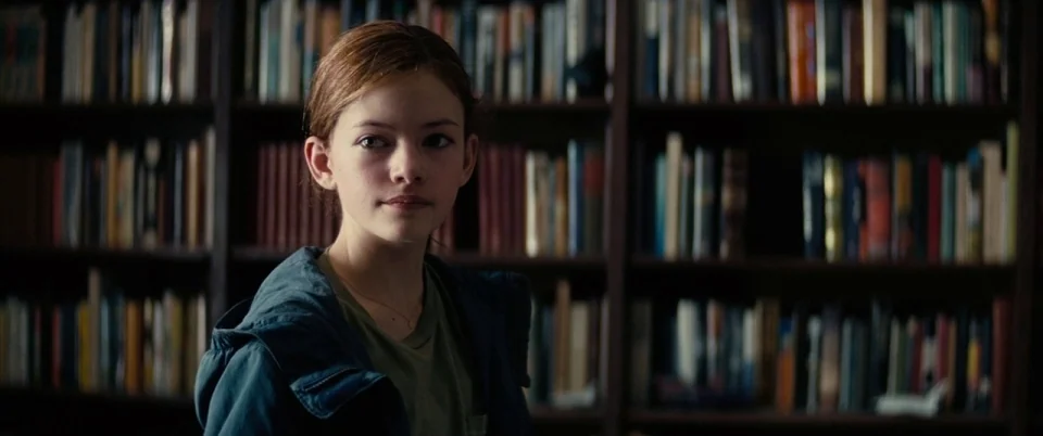 Interstellar (2014) movie still: medium — A dimly lit home library features dark wooden shelves packed with various colorful books. A young teenage…