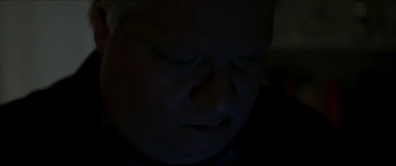 Border movie still: closeup — A close-up shot of a man's face in a darkened environment. He gazes downward with the light source coming from below…