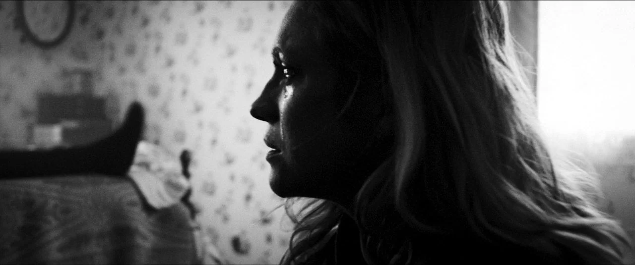 Bushwick (2017) movie still: closeup — A dimly lit room with patterned wallpaper shows a person sitting near a window. A woman is seen in side…