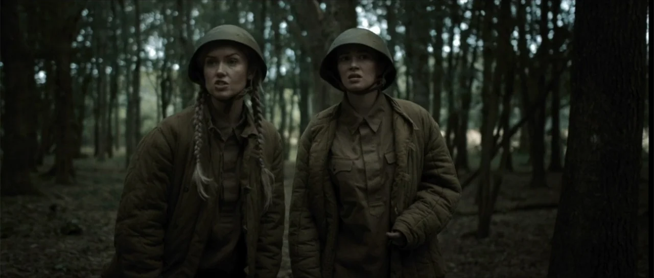 Sin retorno (2009) movie still: medium — A dense forest filled with tall trees and thick undergrowth creates a dark environment. Two female soldiers…