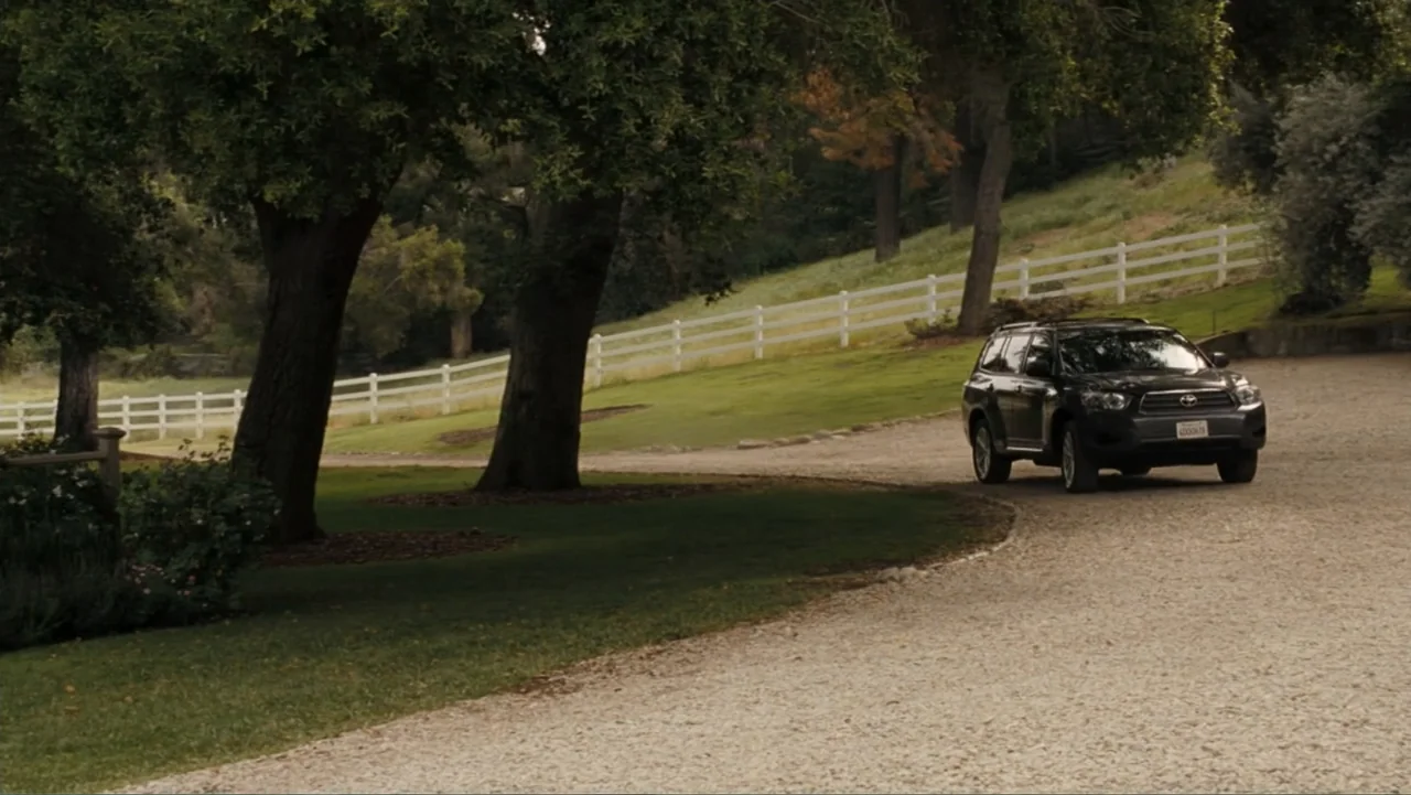 It's Complicated (2009) movie still: wide — A dark SUV is parked on a gravel path adjacent to a large grassy hill. A white wooden fence runs along…