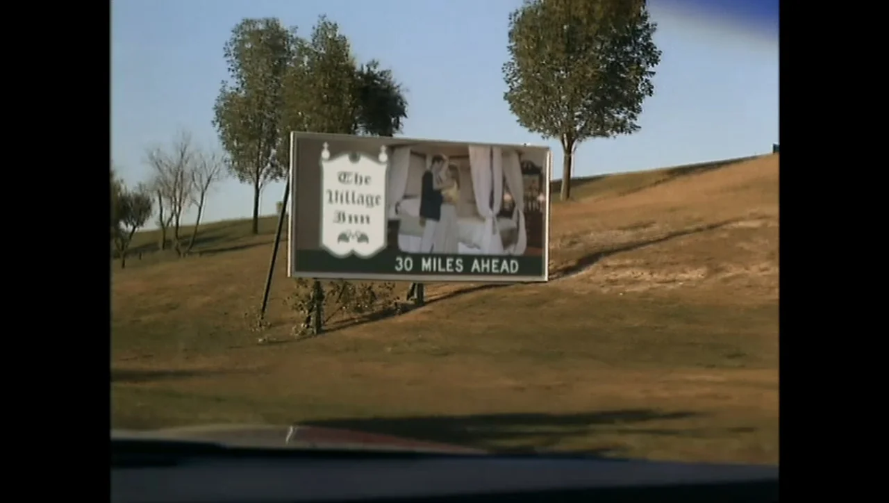 The Awakening (1995) movie still: wide — A rural highway embankment features a rectangular billboard advertising a roadside inn. The sign displays…