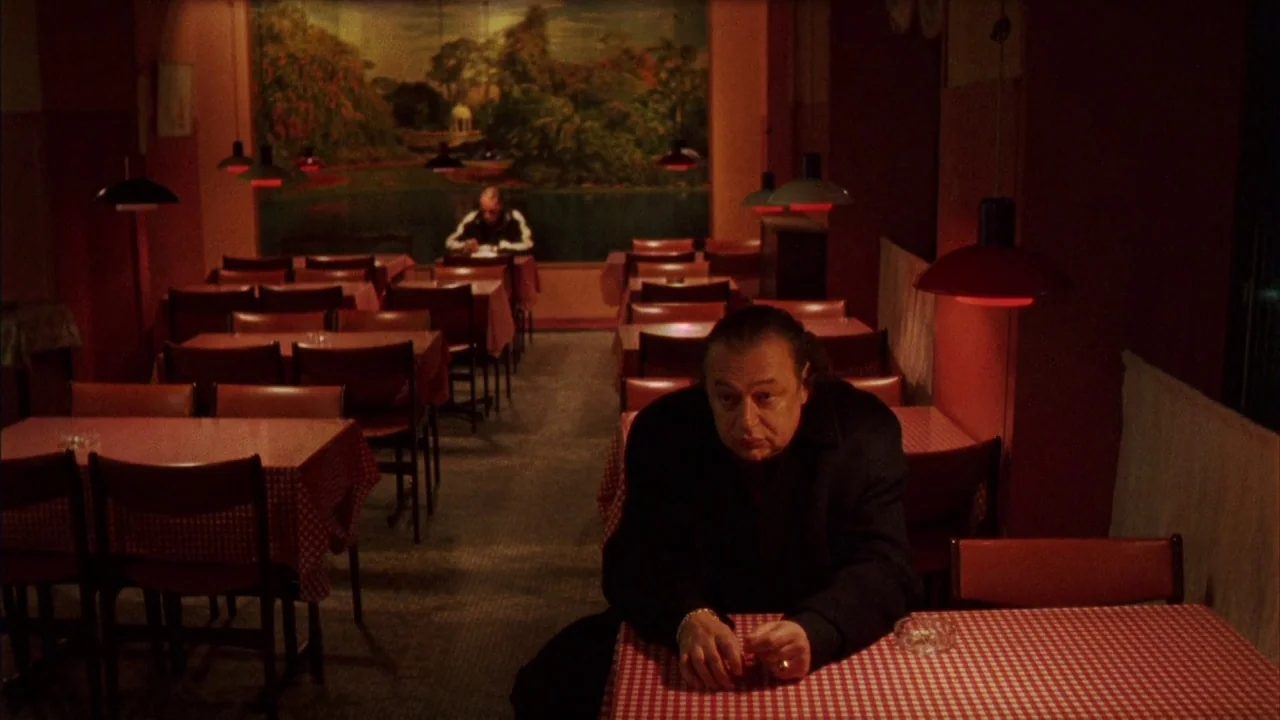 Pusher III (2004) movie still: medium — An dimly lit interior with red checkered tablecloths and red overhead lamps. A man sits at a table in the…