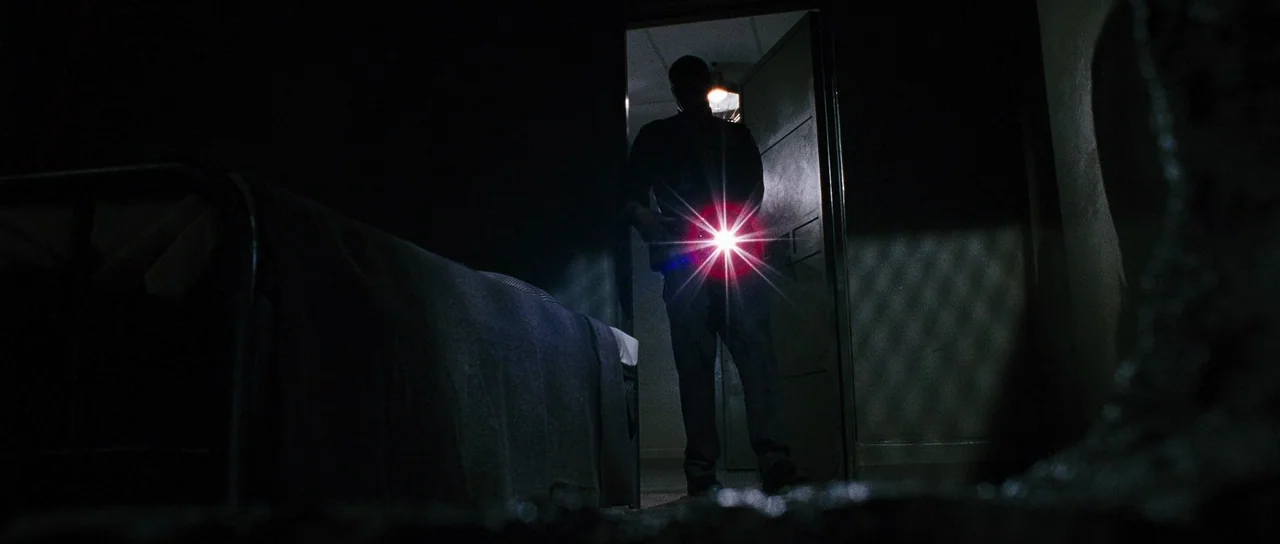 Black Christmas (2006) movie still: medium — A dark room with a metal frame bed in the foreground. A person standing in the doorway shines a bright…