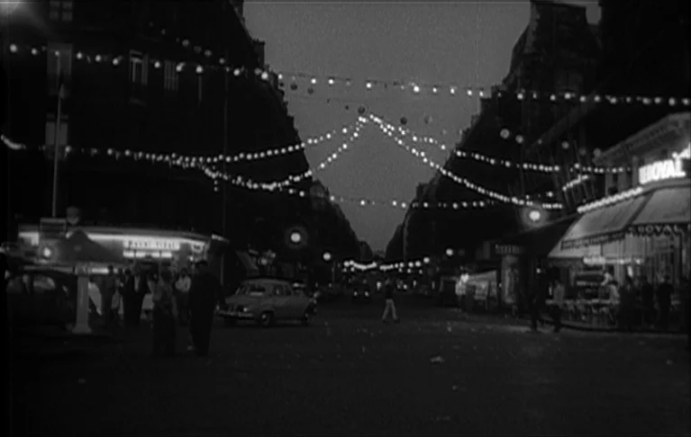 Le Signe du Lion (1962) movie still: wide — A dark urban street at night lined with buildings and glowing string lights. Pedestrians walk on…