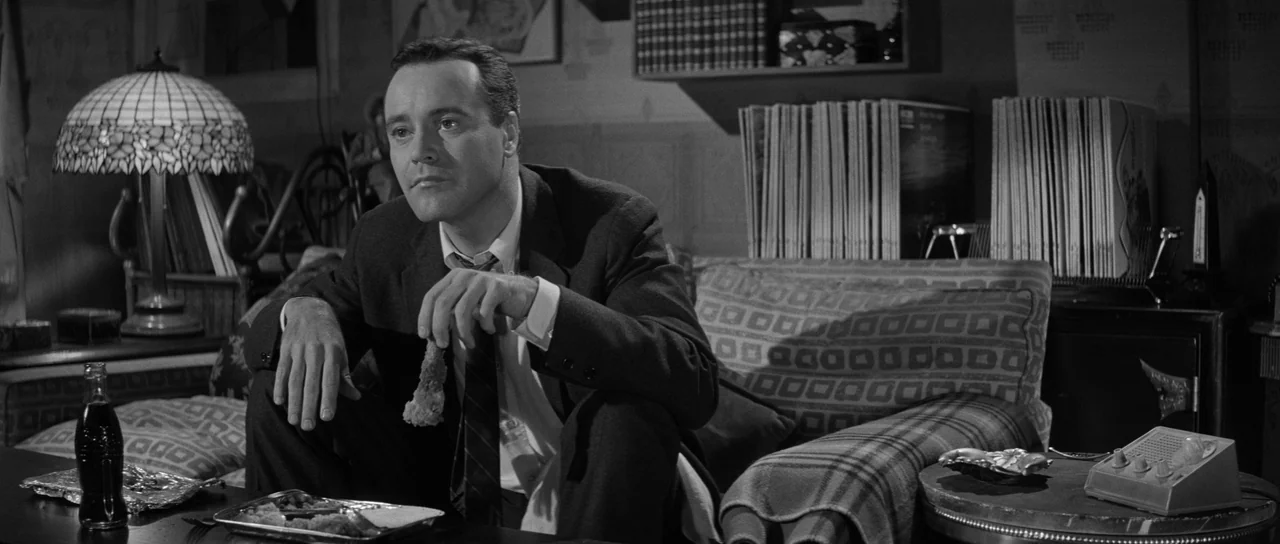 The Apartment (1960) movie still: medium — A 1960s apartment interior is filled with record albums and a patterned couch. A man sits on the couch…