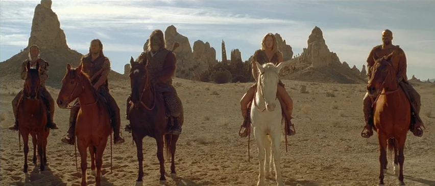 Planet of the Apes (2001) movie still: wide — A group of riders on horses traverses a vast, dry desert with tall stone pinnacles. The figures travel…