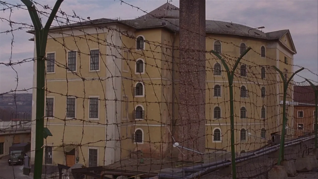 M. Butterfly (1993) movie still: establishing — A multi-story yellow building with a tall brick chimney is enclosed by fencing. Barbed wire crosses…