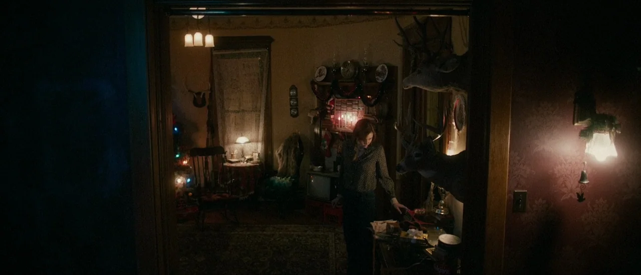 I Trapped The Devil (2019) movie still: wide — A dimly lit 1970s home interior decorated with festive garlands and hunting trophies. A woman stands…