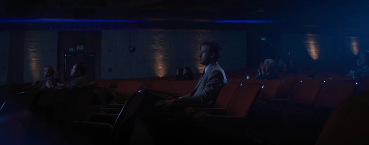 La La Land (2016) movie still: medium — A dimly lit movie auditorium features rows of red velvet seats in darkness. A man sits alone in the…