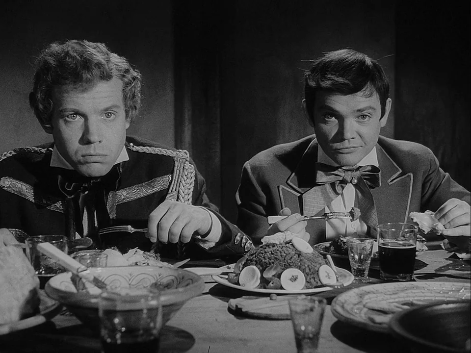 The Magician (1958) movie still: medium — Two men sit at a dark wooden dining table filled with plates and glassware. They look directly at the…