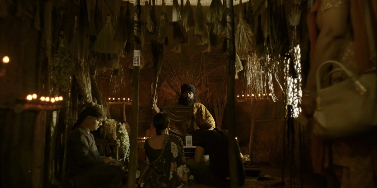 Maja Stoneface (1996) movie still: medium — A dimly lit, rustic hut features a ceiling covered in hanging straw brooms. A man sits center surrounded…
