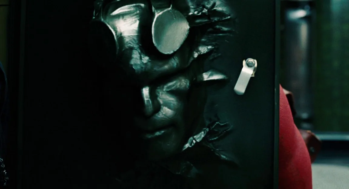 Hellboy II: The Golden Army (2008) movie still: closeup — A dark locker door features a metallic mask sculpted into the surface. The relief includes…