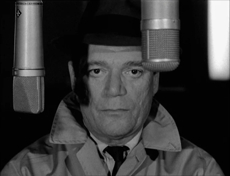 Alphaville (1965) movie still: closeup — A man stands directly behind a professional microphone in a dark room. He stares forward while wearing a…