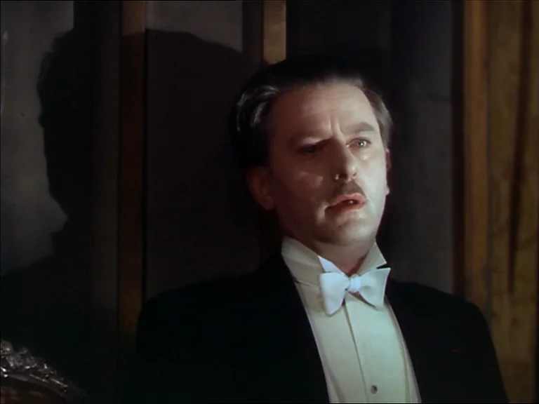 The Red Shoes (1948) movie still: medium — A dimly lit room features a man standing against dark, vertical wooden paneling. He wears a tuxedo and…
